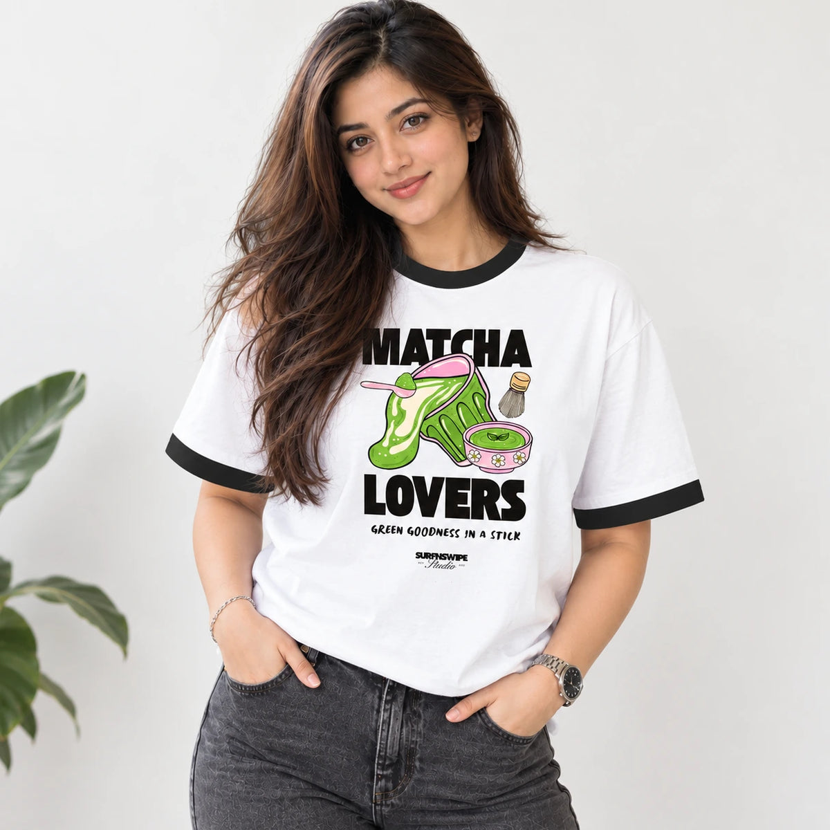 A woman wearing a white ringer t-shirt with black trim and a colorful graphic that reads 'MATCHA LOVERS' with a green shoe illustration, paired with dark gray jeans against a light gray wall.