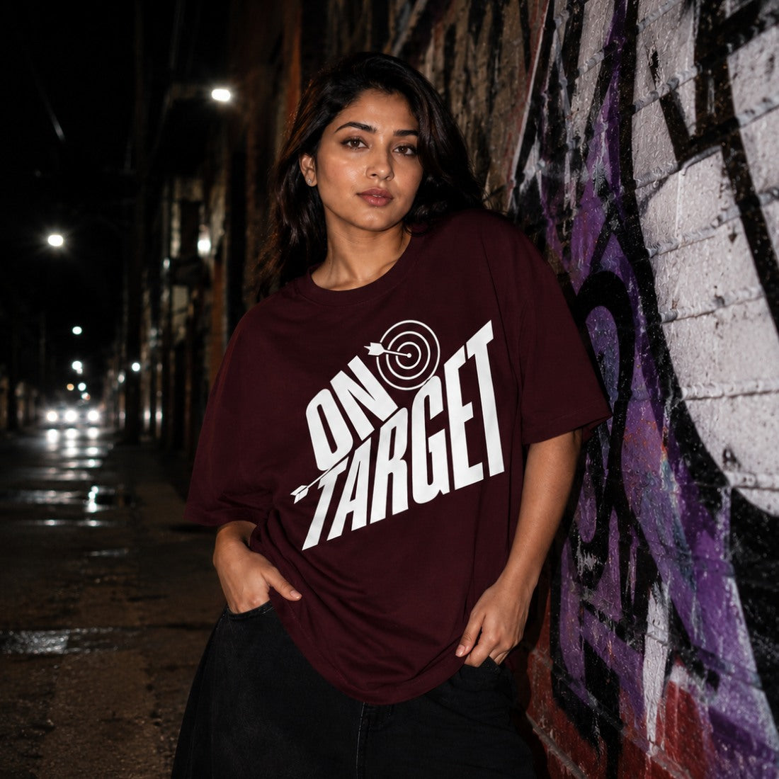 Woman wearing a maroon t-shirt with 'ON TARGET' print against a graffiti-covered wall at night.