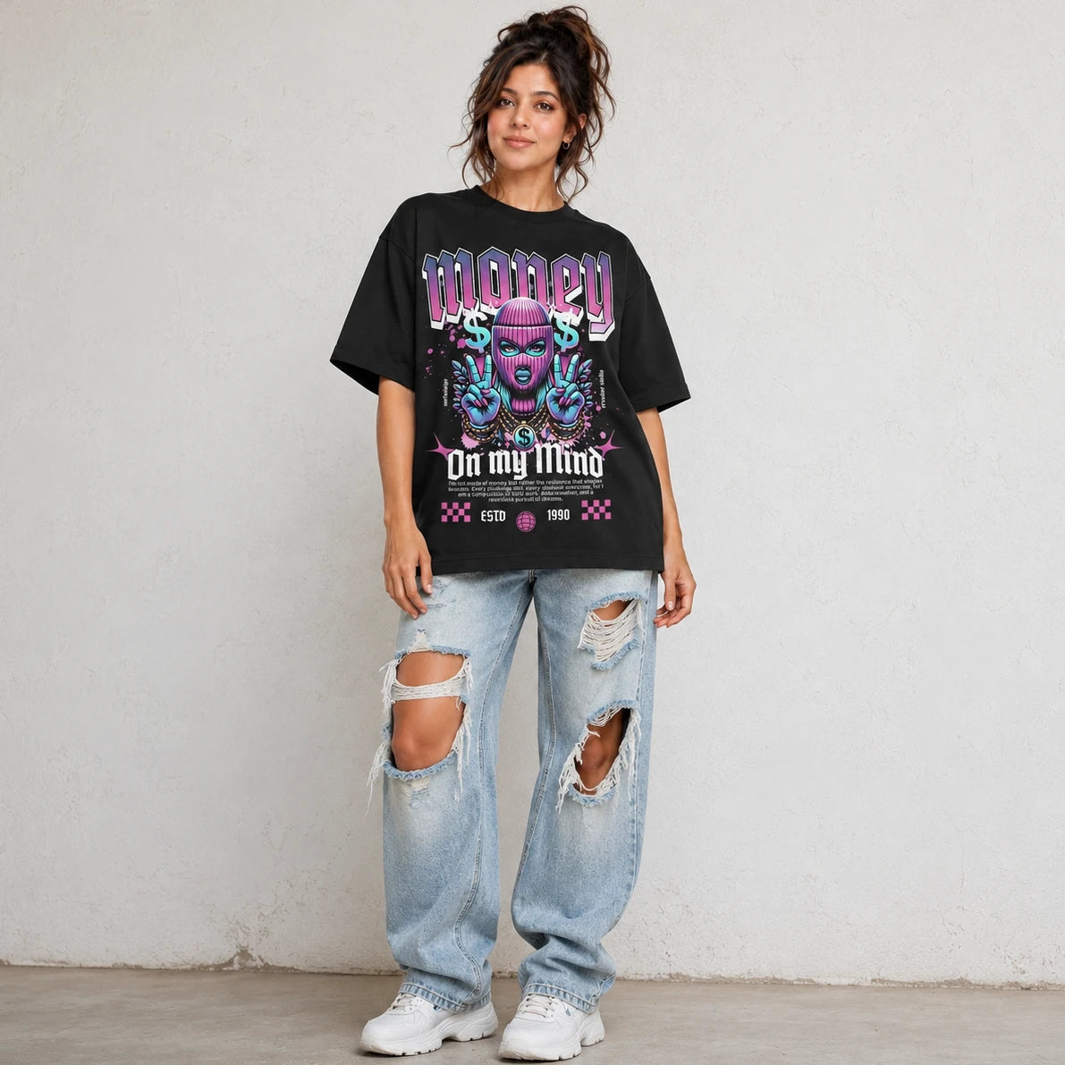 A woman modeling an oversized black graphic t-shirt with a colorful purple and blue design that reads 'MONEY On my Mind', paired with distressed light-wash jeans and white sneakers, standing against a textured off-white wall.