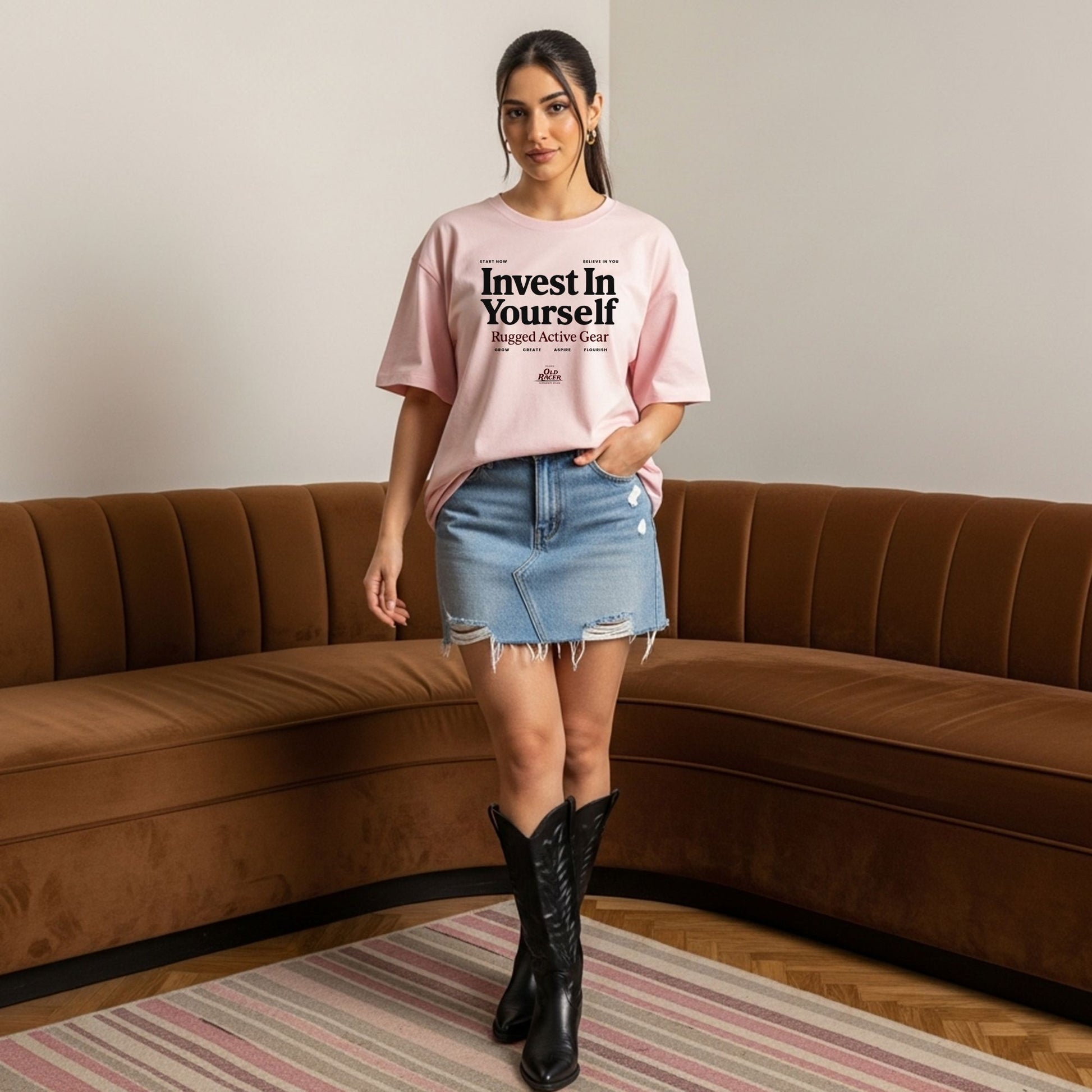 Woman wearing a pink t-shirt with text, denim skirt, and black boots in a living room.