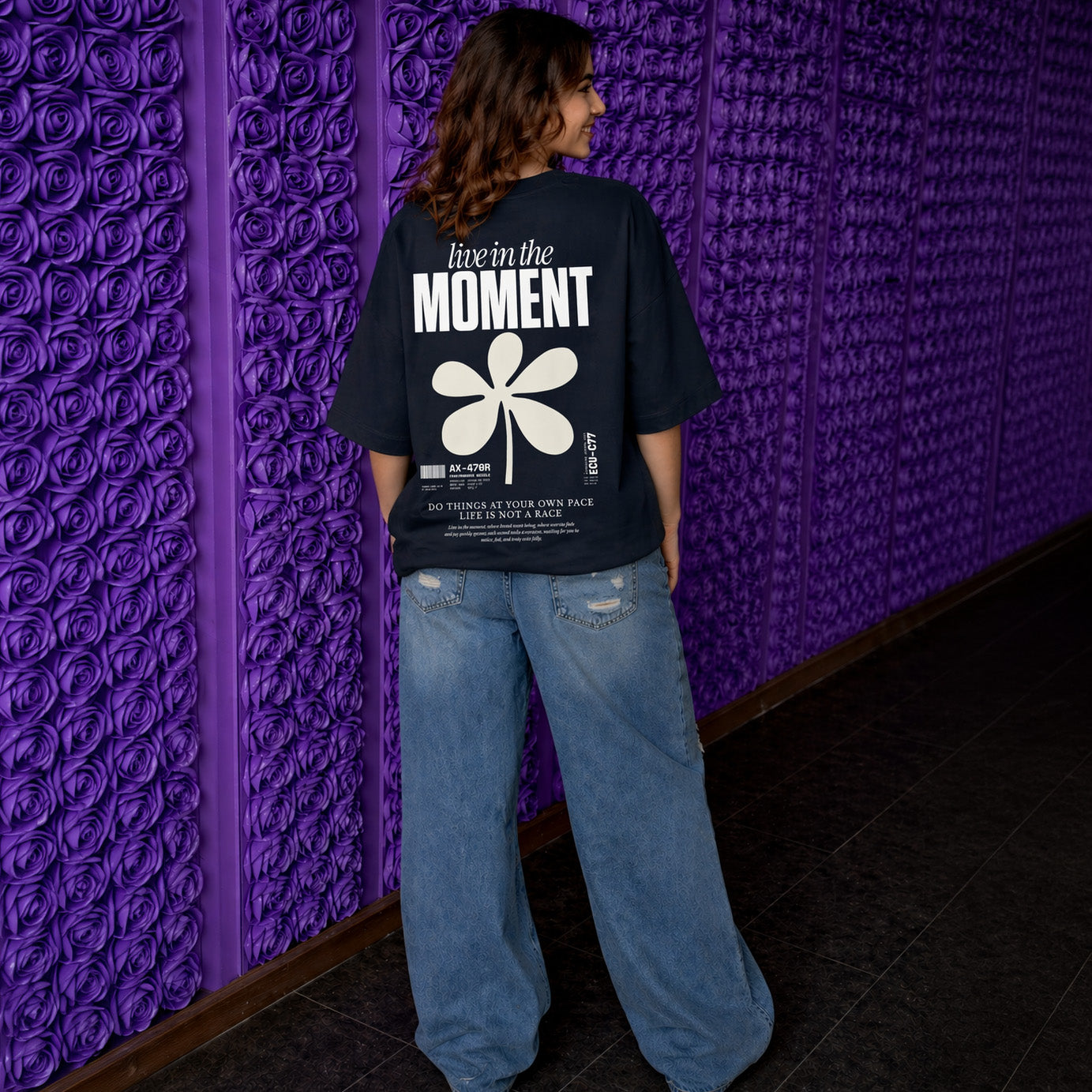 Person wearing a black t-shirt with text and a flower design, standing against a textured purple wall.