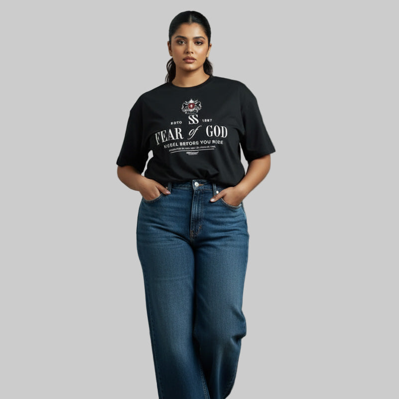 Woman wearing a black 'Fear of God' t-shirt and blue jeans against a gray wall.