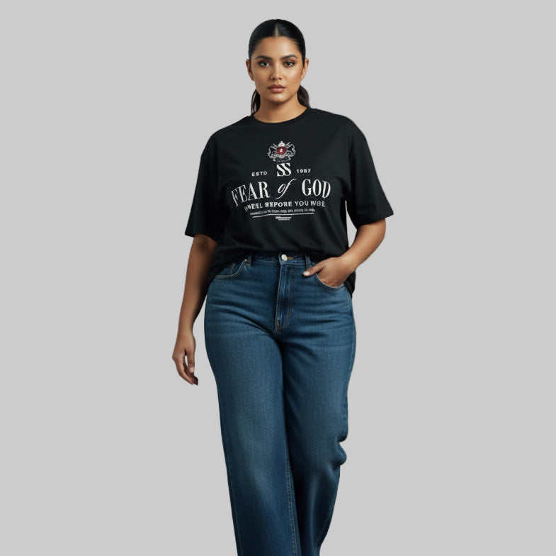 Woman wearing a black 'Fear of God' t-shirt and blue jeans against a gray wall.