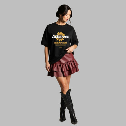 Woman wearing a black t-shirt with 'Achiever' text, burgundy ruffled skirt, and black boots on a neutral background