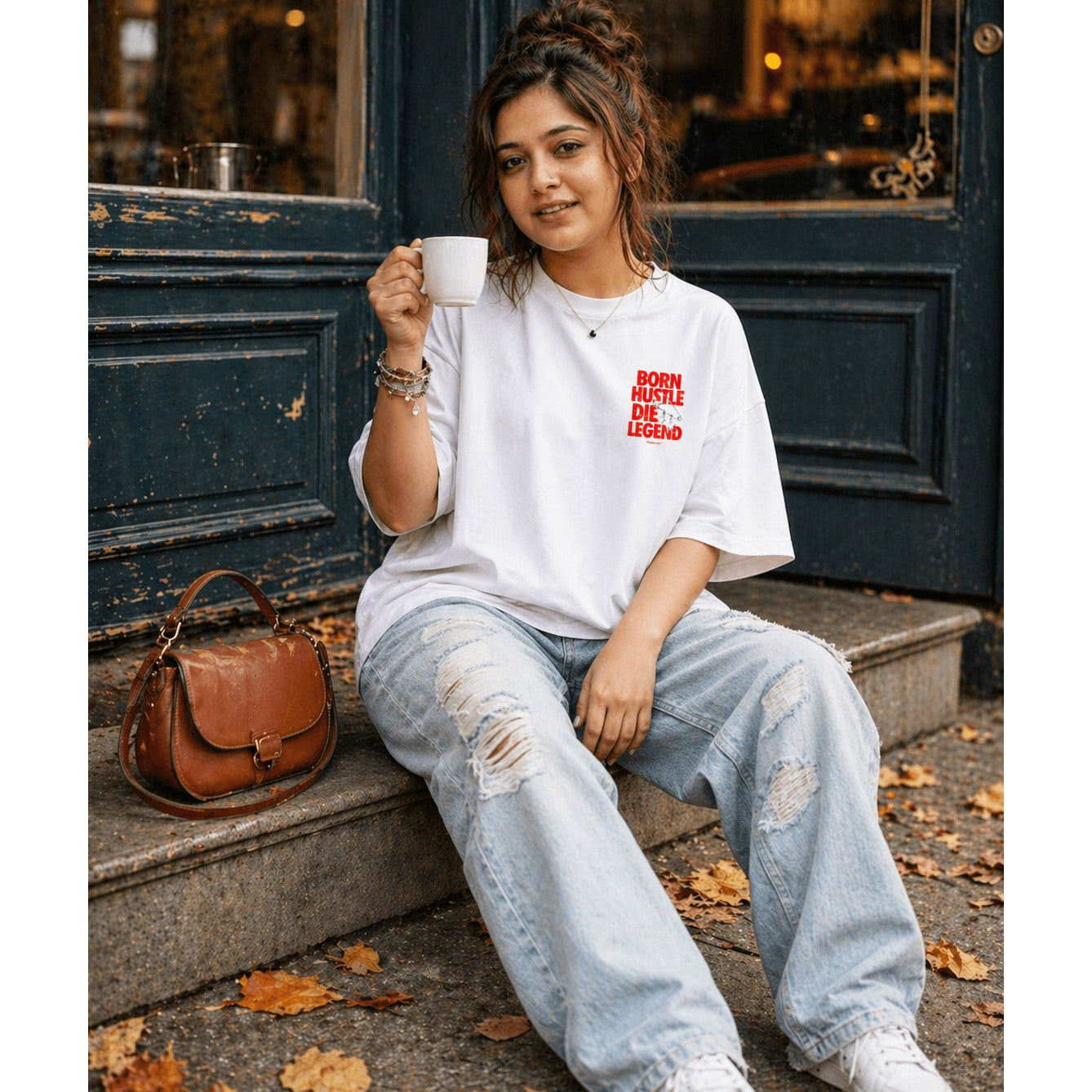 Woman sitting on steps holding a coffee cup, wearing a white t-shirt with red text and ripped jeans.