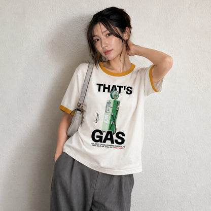 A woman wearing a cream-colored ringer t-shirt with yellow trim and a graphic print that reads 'THAT'S GAS' with a green gas pump illustration. She is wearing gray sweatpants and a light gray shoulder bag, standing against a textured off-white wall.