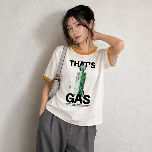 A woman wearing a cream-colored ringer t-shirt with yellow trim and a graphic print that reads 'THAT'S GAS' with a green gas pump illustration. She is wearing gray sweatpants and a light gray shoulder bag, standing against a textured off-white wall.