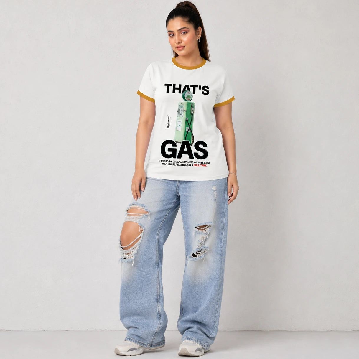 A woman modeling a white ringer t-shirt with yellow trim and a green gas station graphic that reads 'THAT'S GAS'. She is wearing light-wash, distressed wide-leg jeans and white sneakers, standing against a light gray wall.