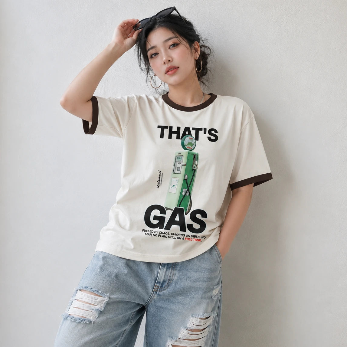 A woman wearing a cream-colored ringer t-shirt with dark brown trim and a green graphic print that reads 'THAT'S GAS'. She is also wearing light-wash, distressed blue jeans, posing against a neutral light gray wall.