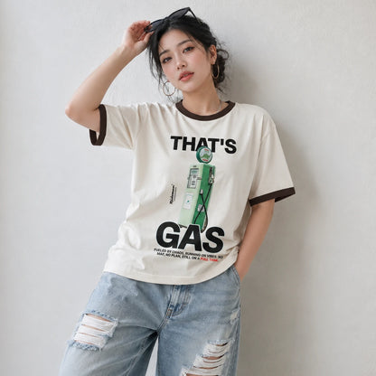 A woman wearing a cream-colored ringer t-shirt with dark brown trim and a green graphic print that reads 'THAT'S GAS'. She is also wearing light-wash, distressed blue jeans, posing against a neutral light gray wall.