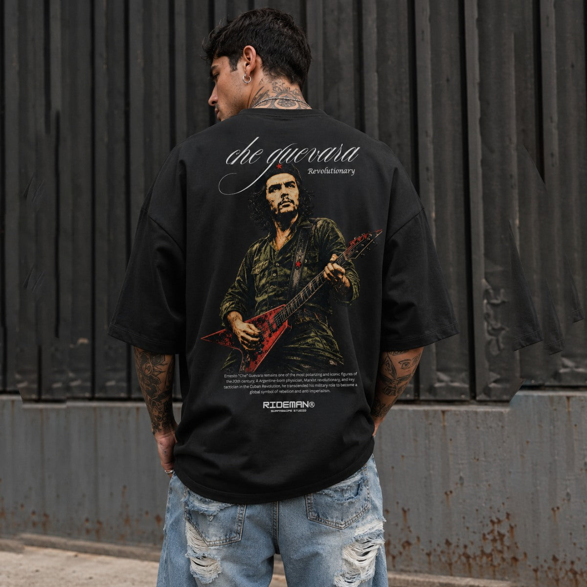 Person wearing a black t-shirt with a graphic design and text, standing against a dark metal wall.