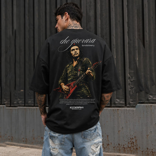 Person wearing a black t-shirt with a graphic design and text, standing against a dark metal wall.