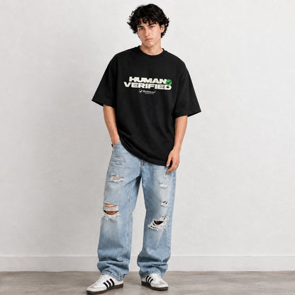 A young man stands against a light gray wall wearing an oversized black graphic T-shirt with 'HUMAN VERIFIED' printed across the chest, light-wash distressed jeans, and white sneakers.