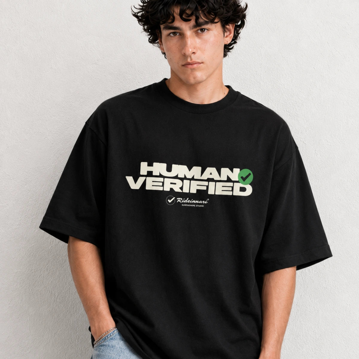A man wearing a black oversized t-shirt with a white and green 'HUMAN VERIFIED' graphic on the chest, posed against a light gray textured wall.