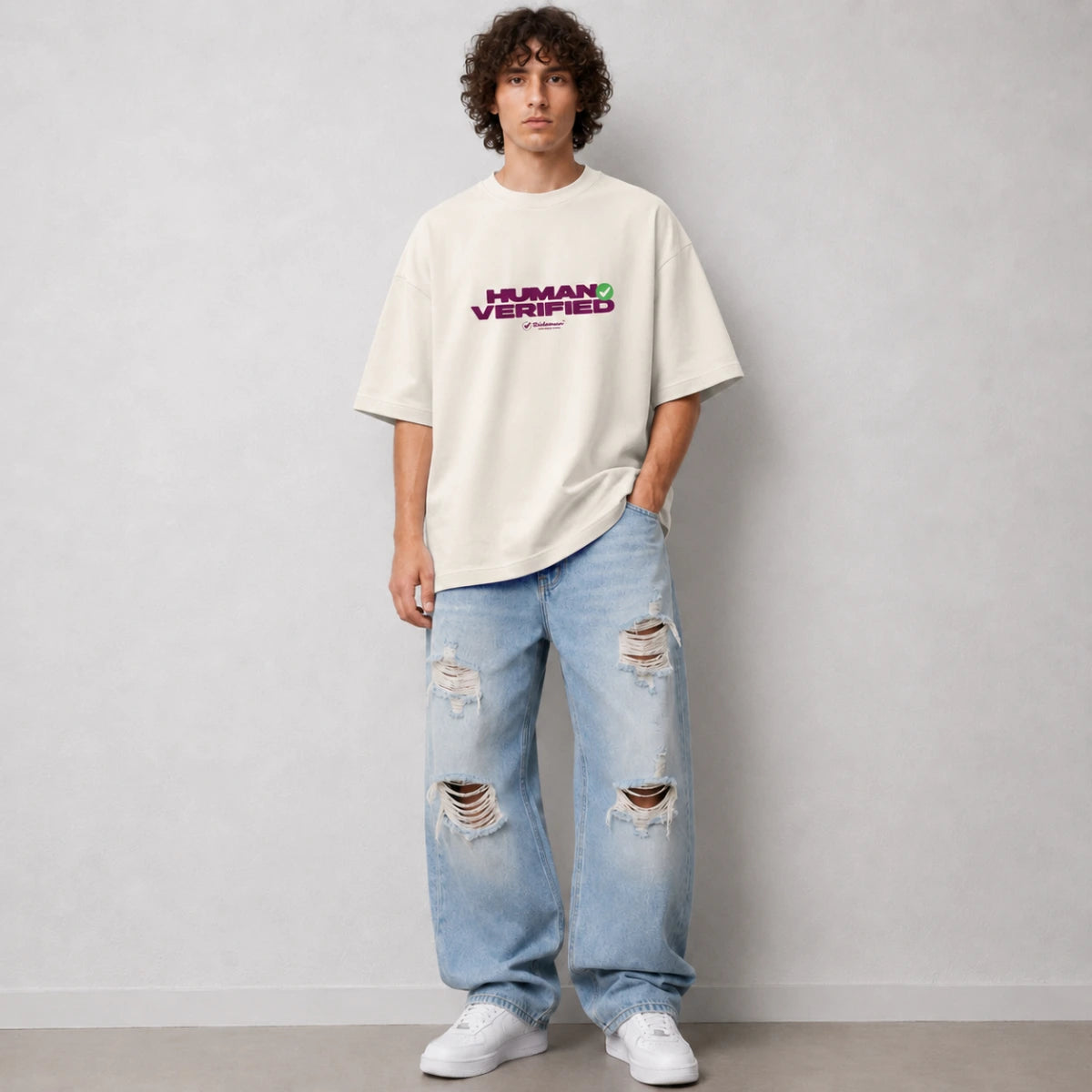 A man wearing an oversized off-white t-shirt with a purple and green 'HUMAN VERIFIED' graphic on the chest, paired with light-wash distressed jeans and white sneakers, standing against a light gray wall.