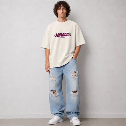 A man wearing an oversized off-white t-shirt with a purple and green 'HUMAN VERIFIED' graphic on the chest, paired with light-wash distressed jeans and white sneakers, standing against a light gray wall.