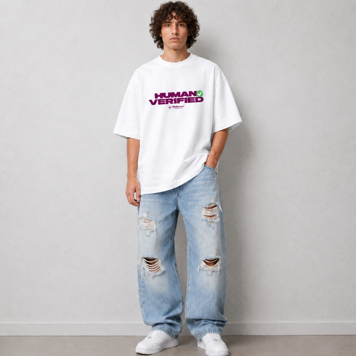 A man wearing an oversized white t-shirt with a purple and green 'HUMAN VERIFIED' graphic, paired with distressed light blue jeans and white sneakers, standing against a light gray wall.
