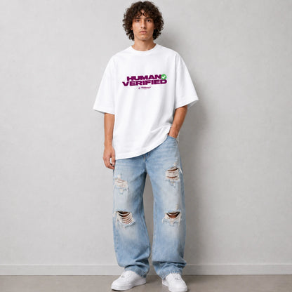 A man wearing an oversized white t-shirt with a purple and green 'HUMAN VERIFIED' graphic, paired with distressed light blue jeans and white sneakers, standing against a light gray wall.
