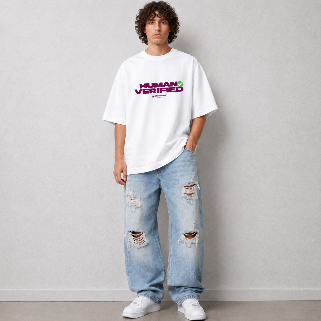 A man wearing a white oversized t-shirt with a purple and green 'HUMAN VERIFIED' graphic, paired with light-wash distressed jeans and white sneakers, standing against a light gray wall.