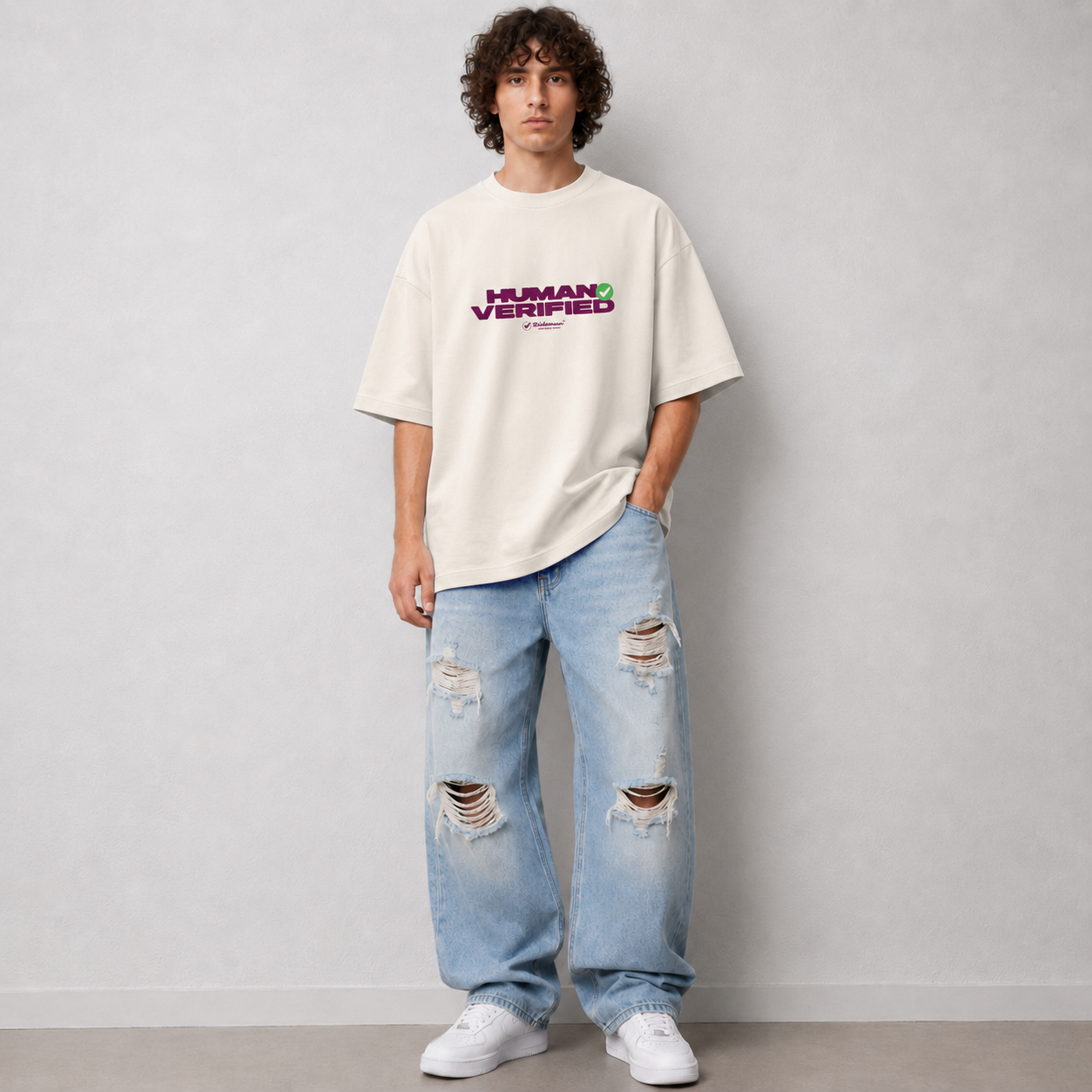 A man wearing an oversized off-white t-shirt with a purple and green 'HUMAN VERIFIED' graphic on the chest, paired with light-wash distressed jeans and white sneakers, standing against a light gray wall.