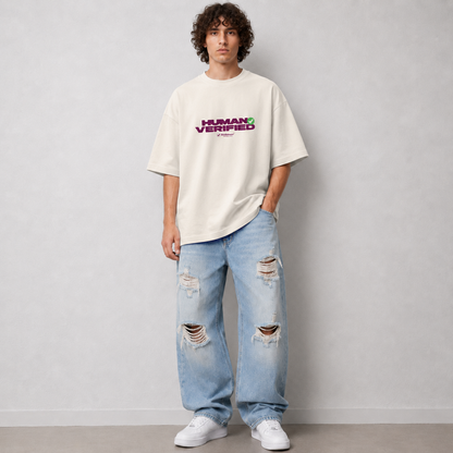 A man wearing an oversized off-white t-shirt with a purple and green 'HUMAN VERIFIED' graphic on the chest, paired with light-wash distressed jeans and white sneakers, standing against a light gray wall.