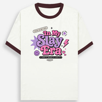 An off-white ringer t-shirt with dark brown ribbed collar and sleeve cuffs, featuring a colorful graphic on the chest that reads 'In My Slay Era' in purple and pink bubble letters with star and lightning accents.