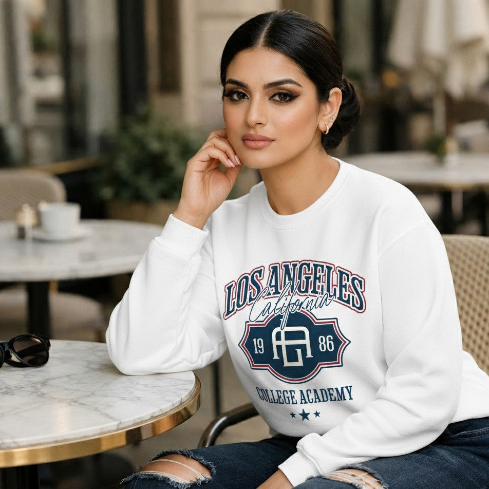 Woman wearing a white sweatshirt with 'Los Angeles College Academy' print, sitting in a cafe.