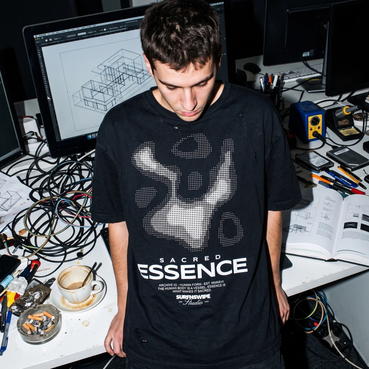 A man wearing a black t-shirt with a white graphic print and the text 'SACRED ESSENCE' stands in a cluttered studio or workshop environment with computer monitors, wires, and tools on a desk.