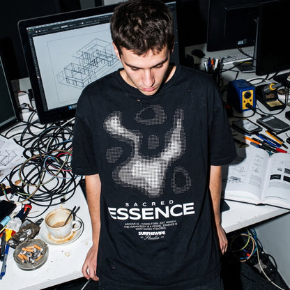A man wearing a black t-shirt with a white graphic print and the text 'SACRED ESSENCE' stands in a cluttered studio or workshop environment with computer monitors, wires, and tools on a desk.