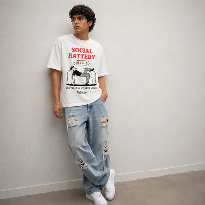 A man wearing a white graphic t-shirt with 'SOCIAL BATTERY' printed in red and black, paired with distressed light blue jeans and white sneakers, leaning against a light gray wall.