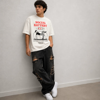 A man wearing a white graphic t-shirt with a red and black print that reads 'SOCIAL BATTERY (E) OUT of SERVICE', paired with distressed black jeans and white sneakers, leaning against a light gray wall.