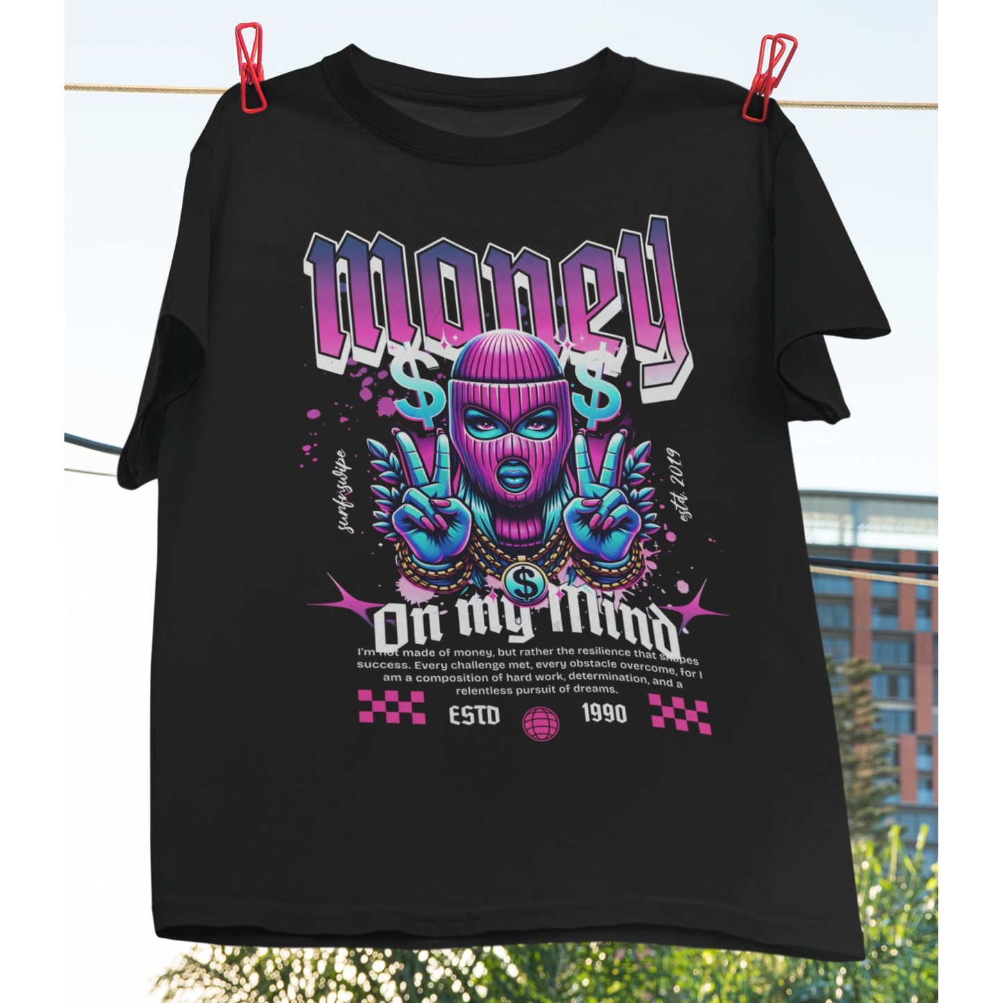 A black oversized t-shirt with a colorful graphic print that includes text and graphics related to money and fashion, displayed on a hanger outdoors.