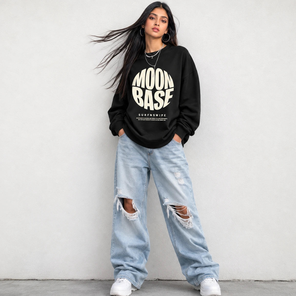 A woman stands against a light gray wall wearing a black sweatshirt with a large cream 'MOON BASE' graphic and distressed light-wash jeans with ripped knees.