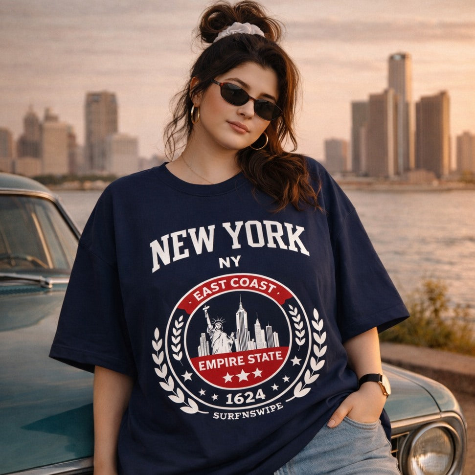Person wearing a navy blue 'New York' t-shirt with a cityscape design, standing by a waterfront with a city skyline in the background.