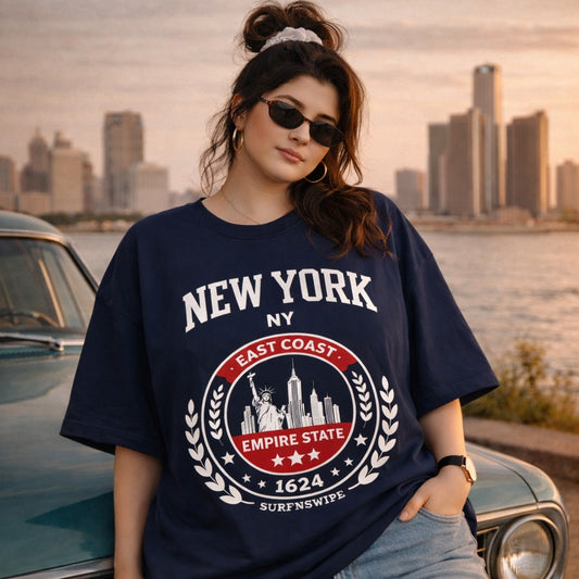 Person wearing a navy blue 'New York' t-shirt with a cityscape design, standing by a waterfront with a city skyline in the background.