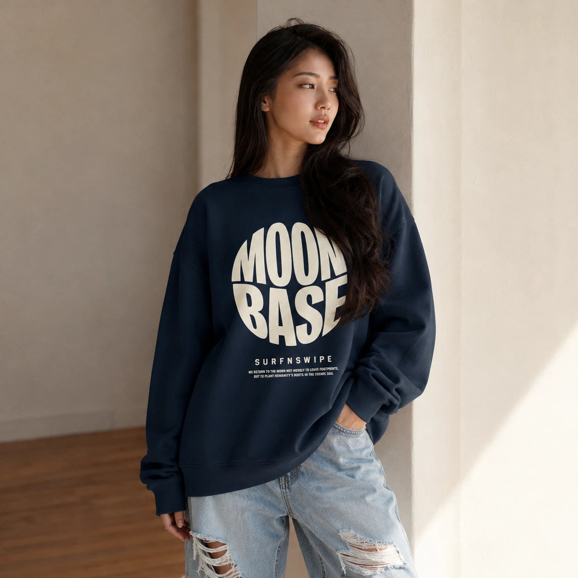 A woman wearing a navy blue crewneck sweatshirt with a large white 'MOON BASE' graphic on the front, paired with light-wash distressed jeans, standing in a minimalist beige-toned room.