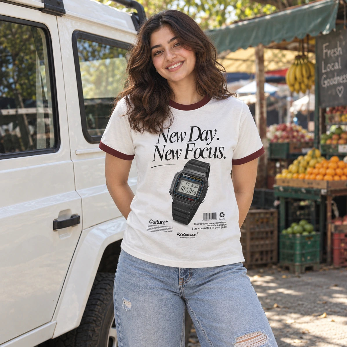 A woman wearing a white ringer t-shirt with dark red trim and a black graphic of a digital watch, standing next to a white vehicle in an outdoor market setting.