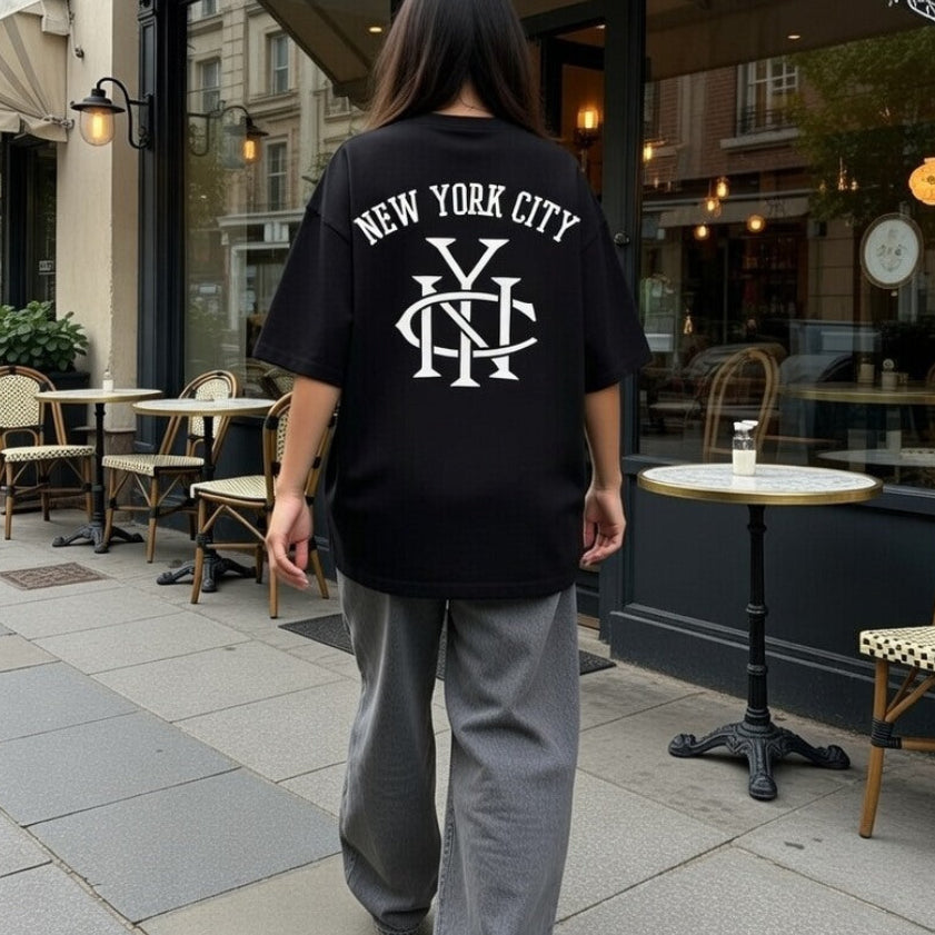 Person wearing a black 'New York City' t-shirt walking on a sidewalk with a cafe in the background.