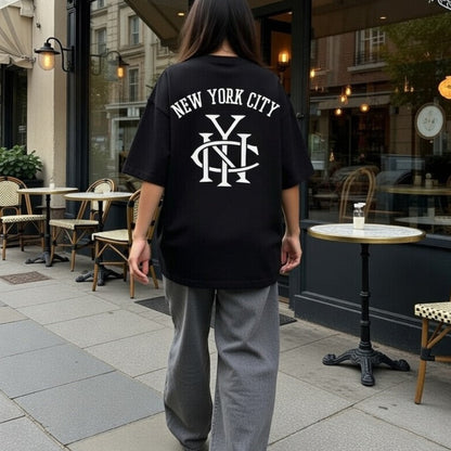 Person wearing a black 'New York City' t-shirt walking on a sidewalk with a cafe in the background.