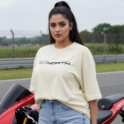 Woman wearing a cream t-shirt with 'True North' text, standing next to a red motorcycle on a track.