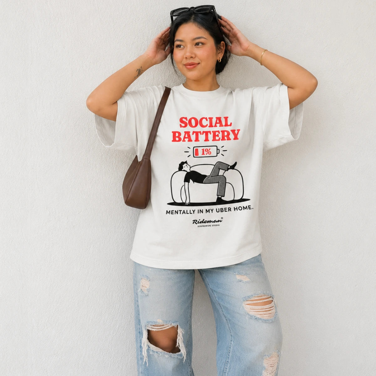 A woman wearing a white graphic t-shirt with a red and black 'SOCIAL BATTERY' design, paired with light blue ripped jeans and a brown shoulder bag, posing against a white wall.