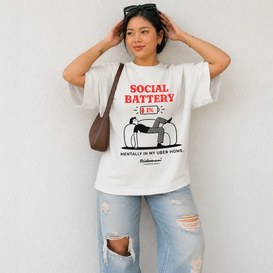 A woman wearing a white graphic t-shirt with a red and black 'SOCIAL BATTERY' design, paired with light blue ripped jeans and a brown shoulder bag, posing against a white wall.