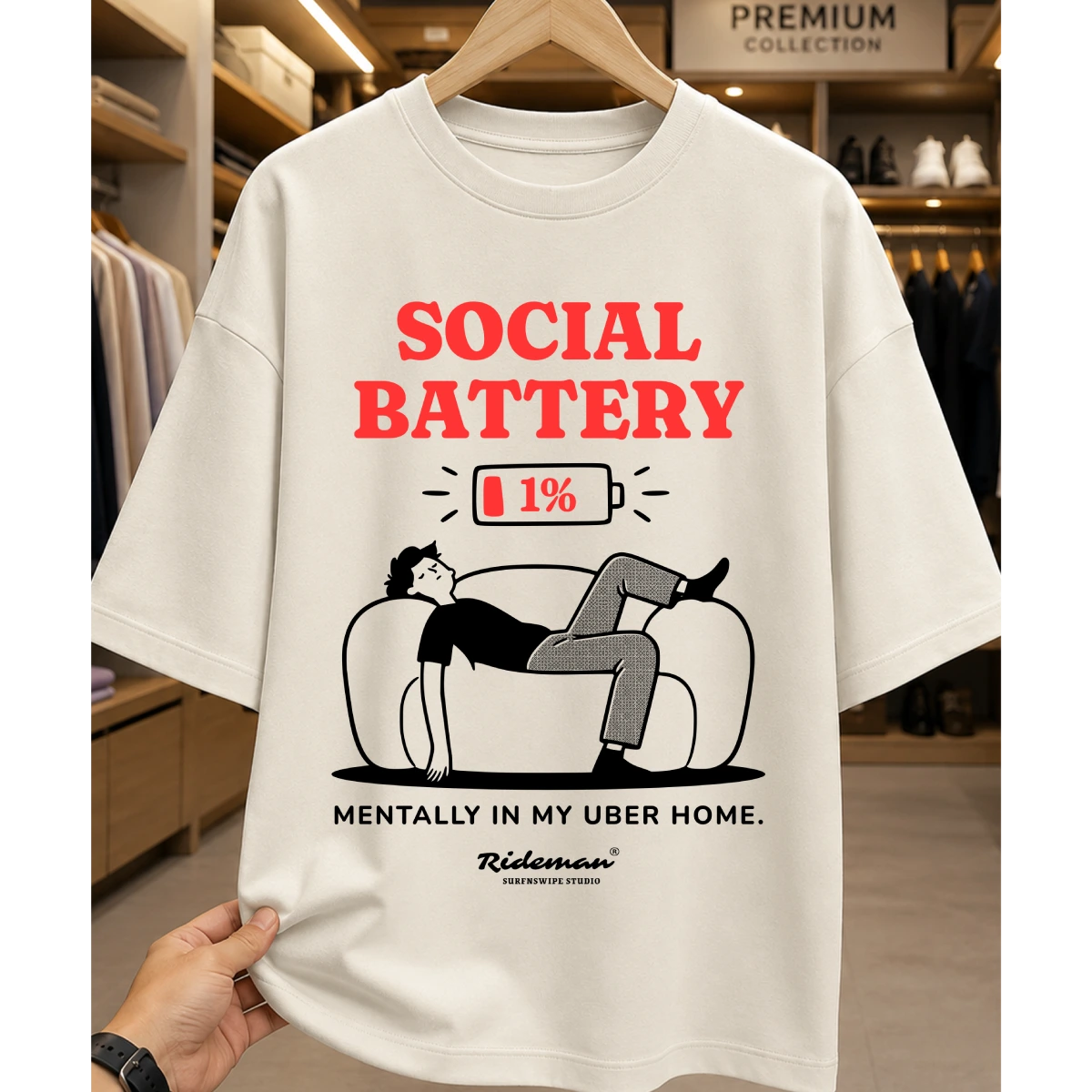 A cream-colored graphic t-shirt hanging on a wooden hanger, held by a hand at the bottom left. The shirt features a humorous graphic with the text 'SOCIAL BATTERY' in red, a 1% battery icon, an illustration of a person on a couch, and the text 'MENTALLY IN MY UBER HOME.