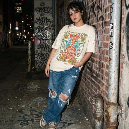 Person wearing a vintage bird print oversized graphic t-shirt and ripped jeans leaning against a graffiti-covered wall.