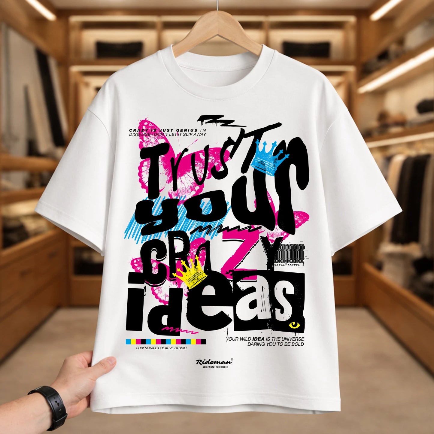 A white short-sleeve graphic T-shirt hanging on a wooden hanger, with a hand holding the bottom left hem. The shirt features a colorful graffiti-style graphic with the words 'Trust your crazy ideas' in black, pink, blue, and yellow.