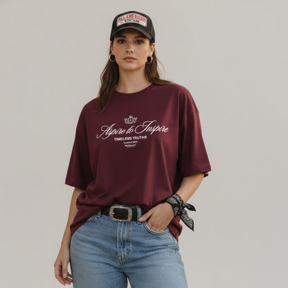 Woman wearing a burgundy t-shirt with text, blue jeans, and a cap on a plain background