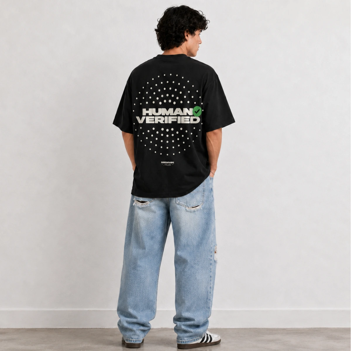 A man stands with his back to the camera wearing a black t-shirt with a white and green 'HUMAN VERIFIED' graphic, paired with light-wash blue jeans and black-and-white sneakers, against a neutral studio backdrop.