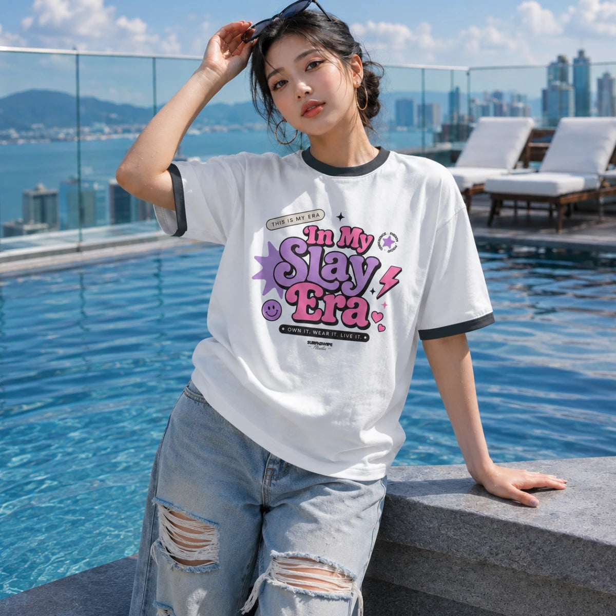 A woman wearing a white ringer T-shirt with black trim and a colorful 'In My Slay Era!' graphic, paired with distressed light blue jeans, posing by a rooftop pool with a city skyline background.