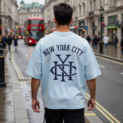Person wearing a light blue 'New York City' t-shirt with a logo, walking on a city street.