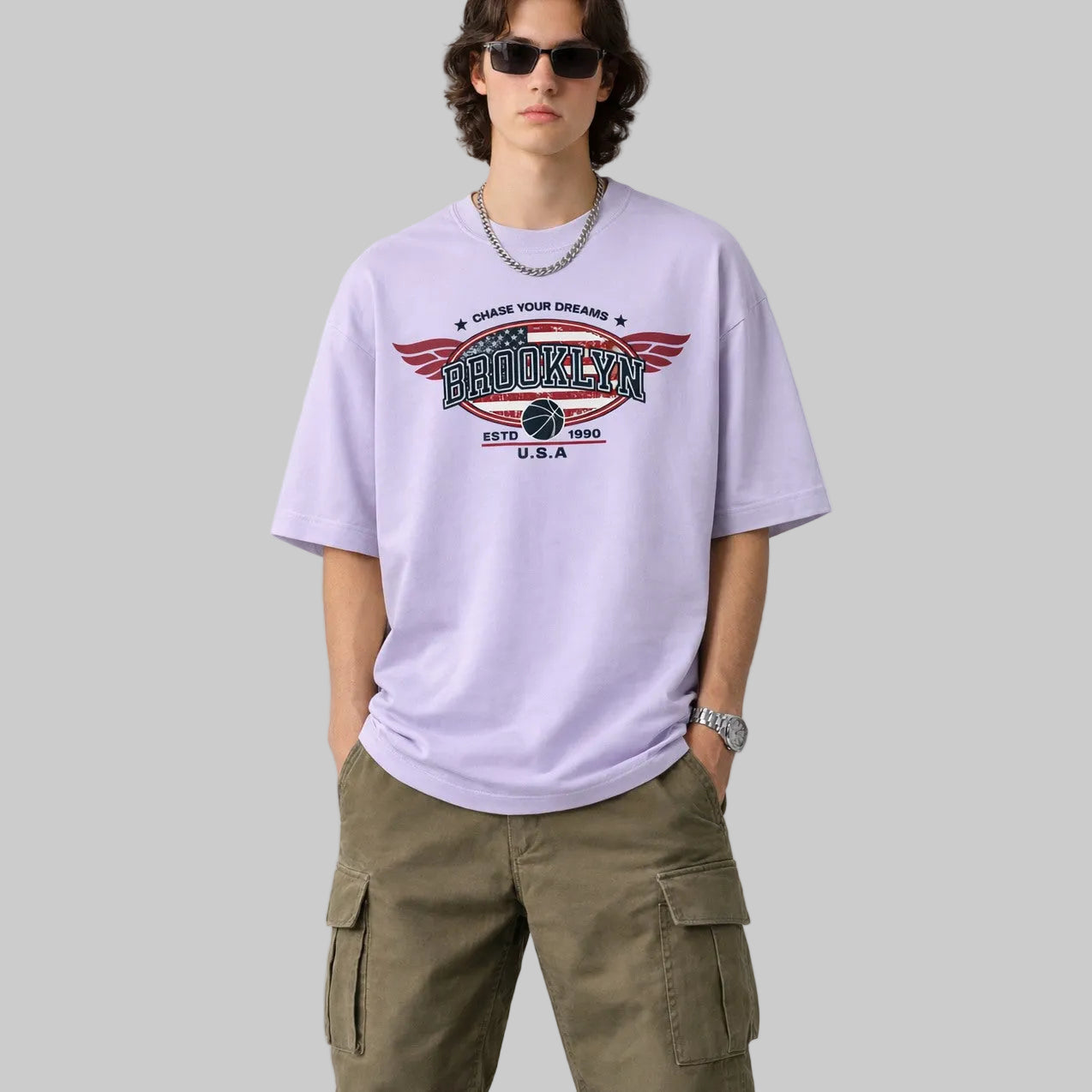 Person wearing a light purple t-shirt with a graphic design and green cargo pants on a plain background